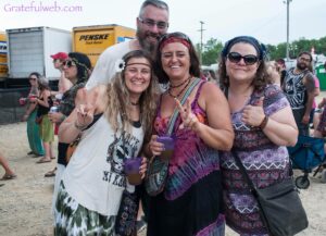 Smiling group posing at an outdoor festival, three women in colorful outfits holding drinks, one man behind them.