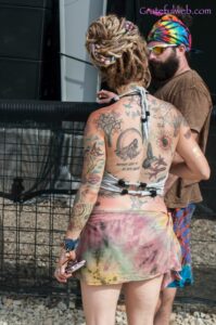 Back view of a topless tattooed woman with colorful dreadlocks and a tie-dye skirt, standing near a fence at an outdoor event, a bearded man with a rainbow headband in the background; watermark 'Gratefulweb.com' visible.