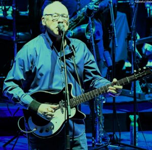 Male musician in blue shirt playing an electric guitar on stage, singing into a mic under blue lighting with a band in the background.