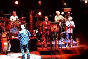 A live band on stage performing with drums, percussion, and microphones under red stage lights; front row saxophonists and a singer visible. A musician in a blue outfit stands with back to camera facing the band.