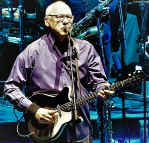 Guitarist wearing glasses sings into a microphone while playing a black electric guitar on stage.