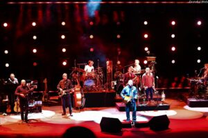 Live band on stage with multiple musicians playing guitars, drums, and keyboard under red stage lights.
