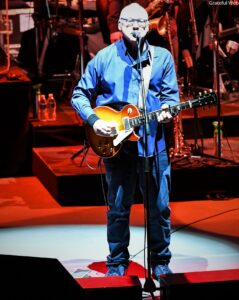 Male guitarist in a blue shirt performs on a lit stage, singing into a microphone with a sunburst electric guitar.