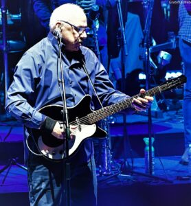 Bald guitarist wearing glasses, playing a black-and-white electric guitar on a blue-lit stage with a microphone in front.