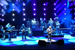 Live band on a blue-lit stage with multiple musicians playing guitars, drums, and keyboards under colorful spotlights.