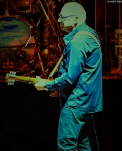 Guitarist in a blue outfit playing an electric guitar on a dim stage with drums in the background.