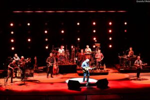 A large band performs on a red-lit stage, with a guitarist in blue at the front center and multiple musicians playing instruments in the background.