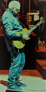Guitarist on stage playing a yellow electric guitar, wearing a blue shirt and jeans with a focused posture on the fretboard