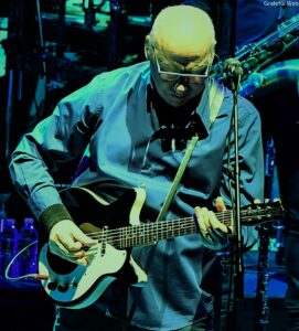 Musician playing an electric guitar on stage, wearing glasses and a blue shirt, microphone nearby, colorful stage lighting in the background