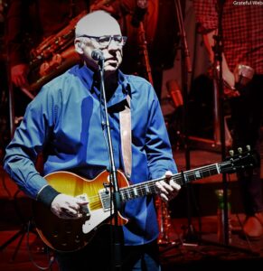 Guitarist in a blue shirt playing a sunburst electric guitar and singing into a microphone on stage.