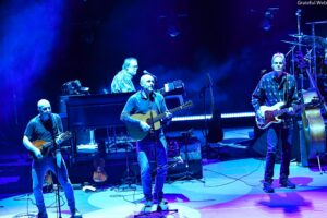 Four musicians perform on a blue-lit stage: two guitars, a keyboard/piano setup, and drums in the background with fog effects.