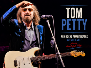 Tom Petty on stage saluting while playing a cream Fender Stratocaster, wearing a blue and black jacket under stage lights.