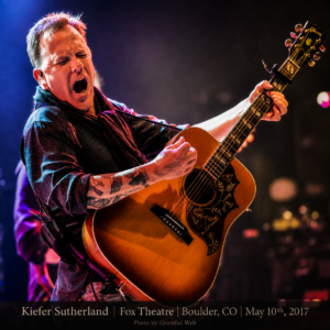 Male guitarist sings passionately on stage, playing an ornate acoustic guitar with eyes closed; bottom caption reads 'Kiefer Sutherland | Fox Theatre | Boulder, CO | May 10th, 2017'