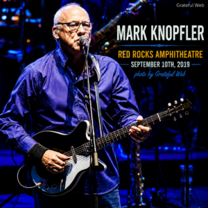 Older male guitarist with glasses performing on a blue-lit stage, singing into a microphone while playing a black electric guitar; concert poster text overlays the right side.