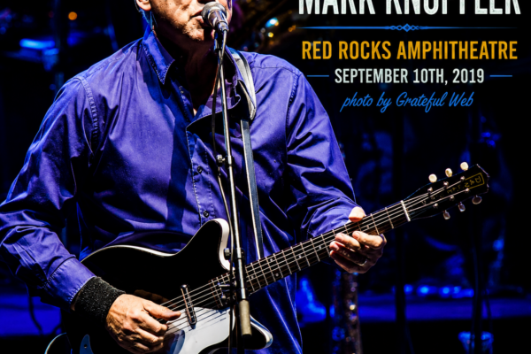 Older male guitarist with glasses performing on a blue-lit stage, singing into a microphone while playing a black electric guitar; concert poster text overlays the right side.