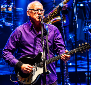 Older man with glasses in a purple shirt sings into a microphone while playing a black-and-white electric guitar on stage with blue lighting.