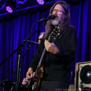 Male guitarist with long hair and beard singing into a microphone on stage under blue/purple lights, wearing glasses and a black shirt.
