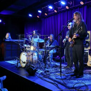 Guitarist with long hair and glasses singing into a mic on a blue-lit stage while a drummer and keyboardist perform nearby.