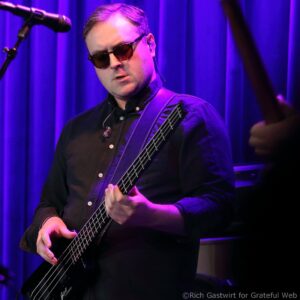 Male bassist wearing sunglasses and a dark shirt, playing a black bass on a purple-lit stage with a microphone visible to the left.