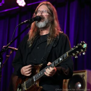 Male guitarist with long hair and beard singing into a microphone on stage, playing a brown electric guitar under purple stage lights.