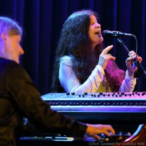 Female singer with long dark hair passionately singing into a microphone on stage, blue-purple lighting illuminating her face and a keyboard in the foreground.