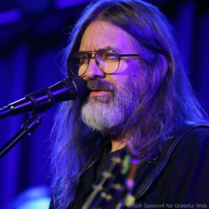 Close-up of a bearded musician with long hair and glasses singing into a microphone on a blue-lit stage.