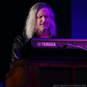 Close-up of a long-haired keyboardist on stage, eyes closed, playing a Yamaha keyboard under purple lighting.