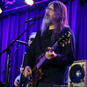 Male guitarist with long gray hair and beard singing into a microphone on a purple-lit stage.