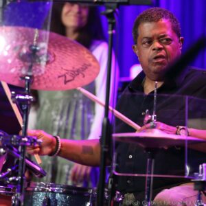 Drummer at a concert, focusing on his face as purple stage lights glow while he plays the drums.