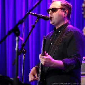 Male musician sings into a microphone while playing an electric guitar on a purple-lit stage.