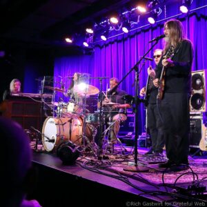 Guitarist with long hair singing into a microphone while playing an electric guitar on a purple-lit stage; drummer in the back.
