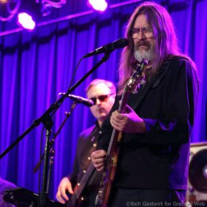 Guitarist with long gray hair and beard playing a guitar on a purple-lit stage, wearing glasses and a black shirt.