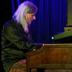 Side profile of a pianist with long silver hair playing a keyboard on a blue-lit stage on dark clothing.