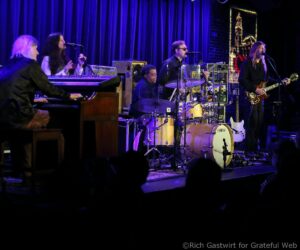 Live band performing on stage with keyboardist, vocalists, drummer, and guitarist under blue/purple lights.