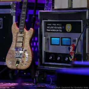 Electric guitar on a stand next to a rack-mounted amplifier, bathed in purple stage lighting at a backstage or stage setup.