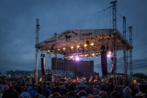 Outdoor concert stage at dusk with colorful lights and a large crowd; banner reads The Stone Pony Stage with 'Summer' on the side doors up.
