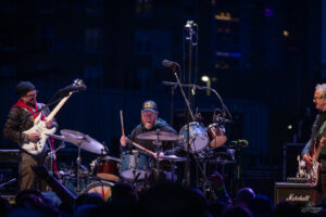 Band performing on a nighttime stage: guitarist left, drummer mid, bassist right, with audience in foreground under blue lighting