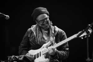 Guitarist wearing a beanie and glasses plays a white electric guitar on stage, smiling at the audience.",