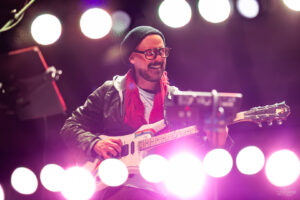 Musician playing an electric guitar on stage under pink-purple concert lights, wearing a black beanie, glasses, and a red scarf.