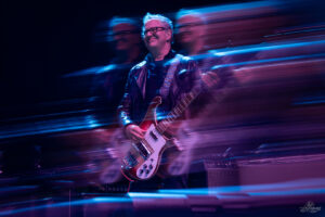 Guitarist in a leather jacket playing a red bass on stage amid blue motion blur and lights.