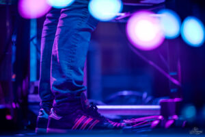 Person's legs in jeans and Adidas Samba sneakers on a pedalboard, stage lighting in purple and blue hues behind.
