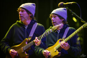 Two men in gray beanies and dark hoodies play matching wooden electric guitars on a dimly lit stage.