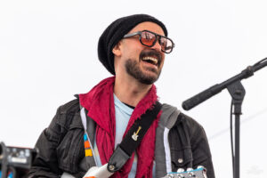 Smiling man wearing a black beanie and sunglasses performs with an electric guitar on a stage with a microphone nearby.