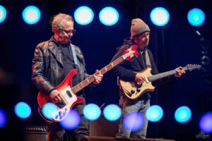 Two male musicians on stage; left plays a red bass guitar, right plays a cream electric guitar, blue stage lights overhead.