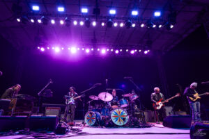 A six-member band performing on a purple-lit stage with drums, guitars, and keyboards, colorful drum faces in the foreground.