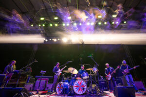 Live band performing on an outdoor stage at night with colorful stage lights; guitarist, bassist, keyboardist, drummer, and percussion setup visible.