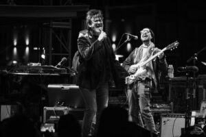 Black-and-white photo of a male singer with glasses performing into a handheld mic while a guitarist with a white electric guitar plays in the background on a concert stage surrounded by equipment and cables.