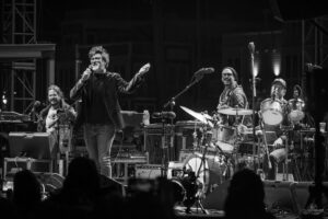 Lead singer in a black jacket performs into a microphone while two bandmates play guitar and drums on a dark stage.