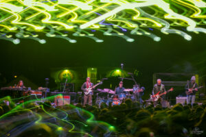 Band performs on a dark stage under swirling neon green and yellow light trails above the crowd.