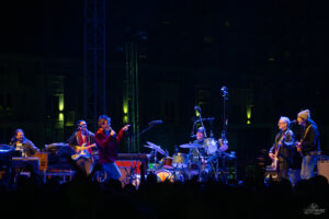 A live band performs on a night-stage with blue and green lighting; musicians play guitars, drums, and keyboards while a singerpoints into the microphone, and an audience silhouette is visible in the foreground.
